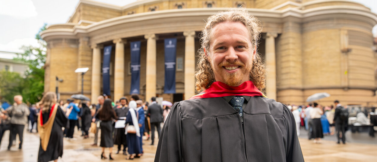 PharmD for Pharmacists Graduate Brandon Toner photographed in graduation regalia with University of Toronto's Convocation Hall in background