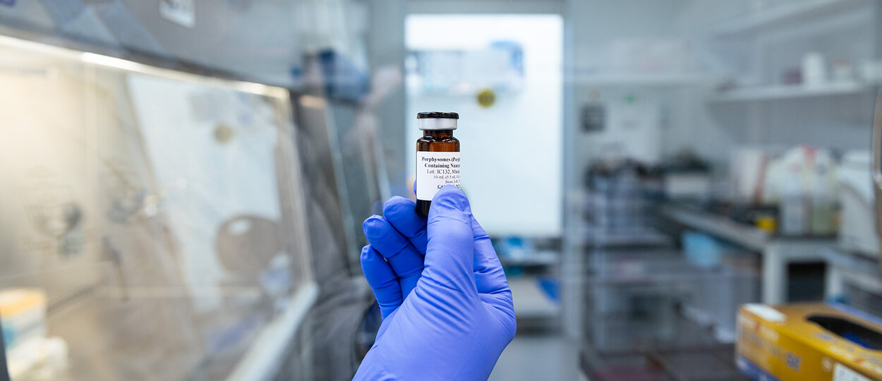 A blue latex glove holding a vial of porphysomes in front of a window into a GMP facility