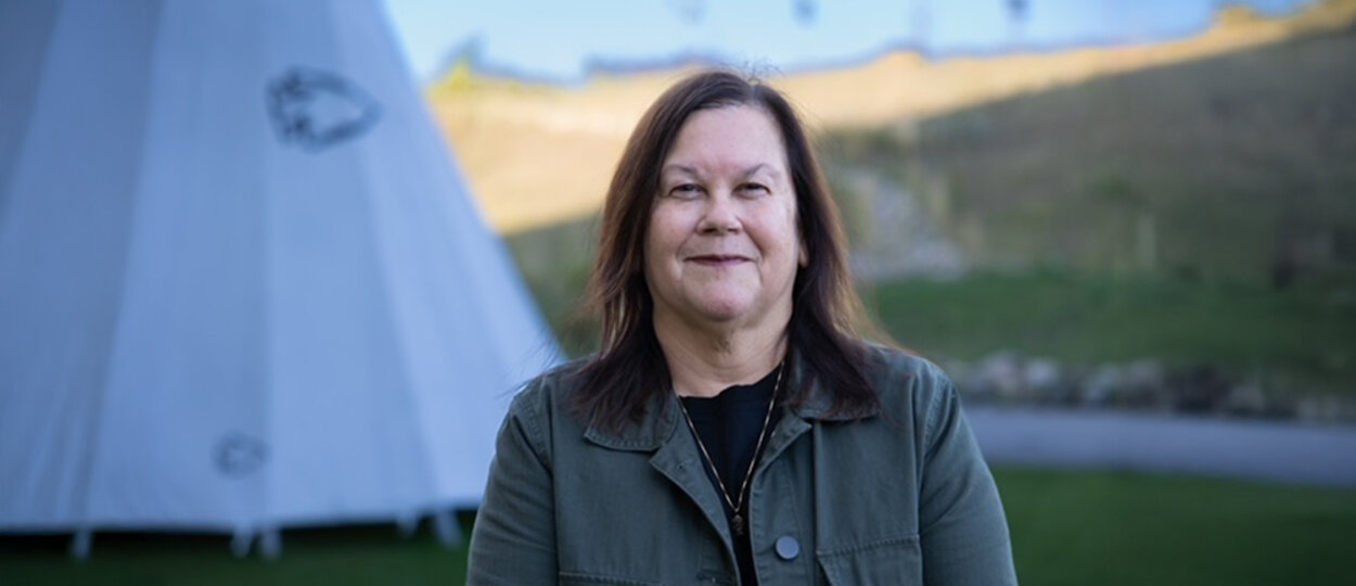 Portrait of a woman standing outdoors with a grassy hill, tipi, and blue sky in the background