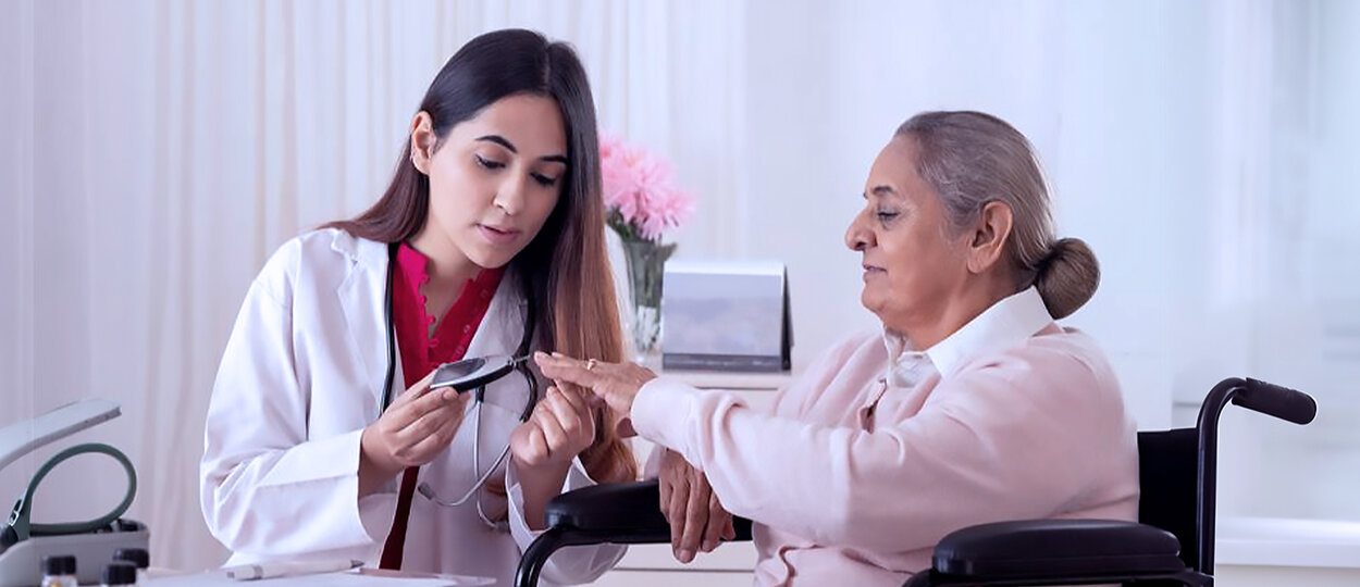 A young healthcare professional checks the blood sugar level of an older woman using a glucometer. The older woman, seated in a wheelchair, extends her hand while the professional focuses attentively on the reading. They are in a bright clinical setting with soft decor, including pink flowers in the background.