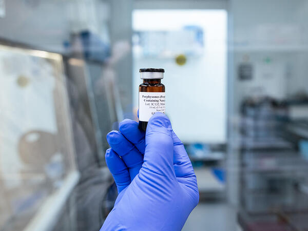 A blue latex glove holding a vial of porphysomes in front of a window into a GMP facility A blue latex glove holding a vial of porphysomes in front of a window into a GMP facility